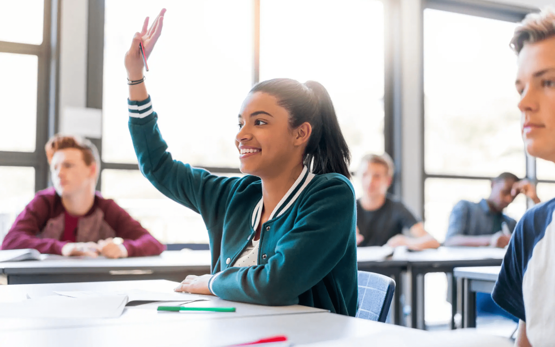 Student raising hand in classroom, smiling.