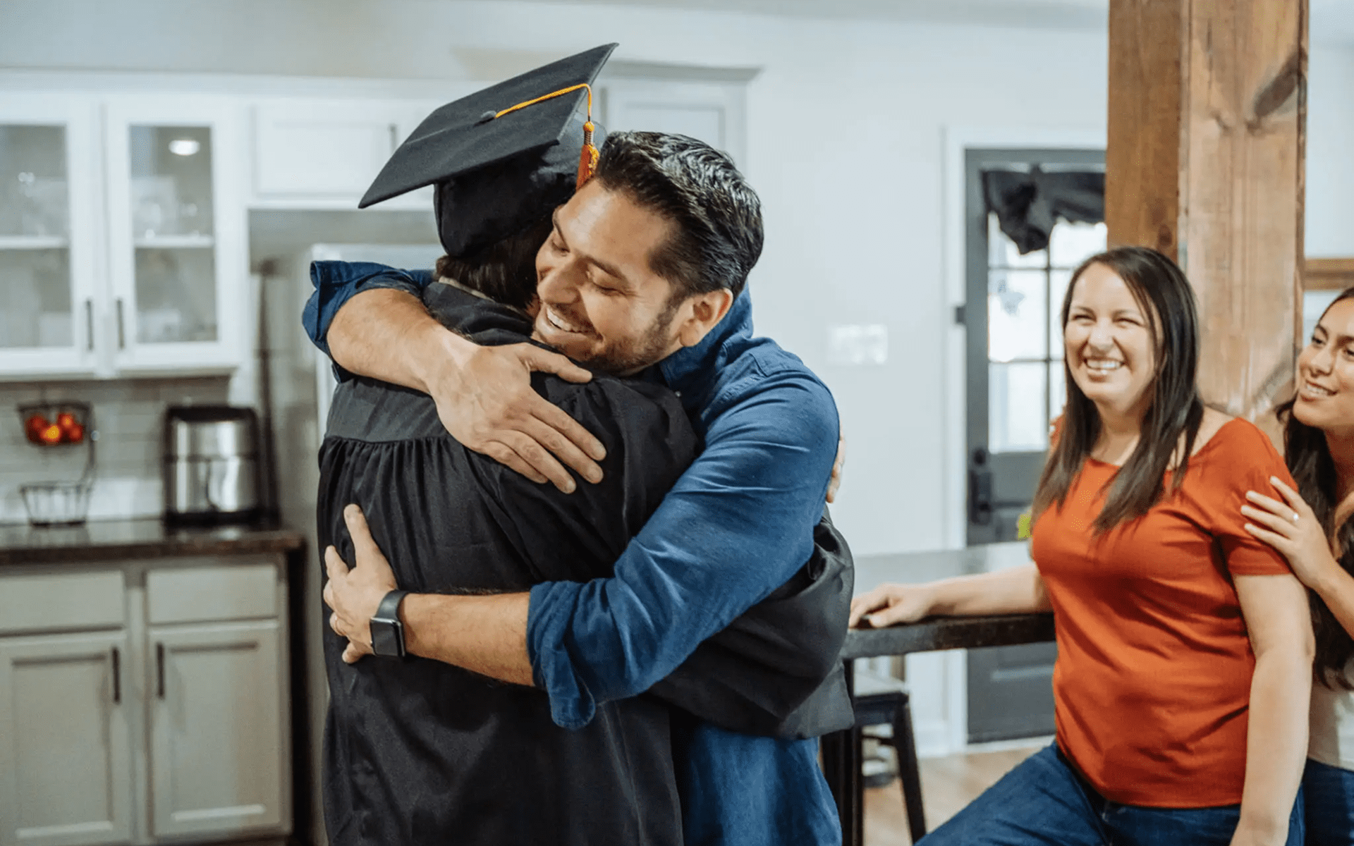 Graduate hugging family in a kitchen celebration.